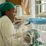 Female health worker caring for a newborn in a neonatal unit beside an incubator and monitoring equipment.