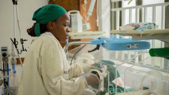 Female health worker caring for a newborn in a neonatal unit beside an incubator and monitoring equipment.